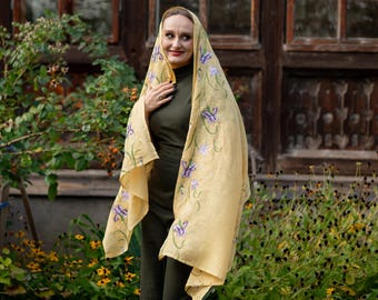 May include: A woman wearing a pale yellow shawl with purple floral embroidery. The shawl is draped over the head and shoulders. The woman is wearing a dark green dress. The background includes a wooden door and greenery.