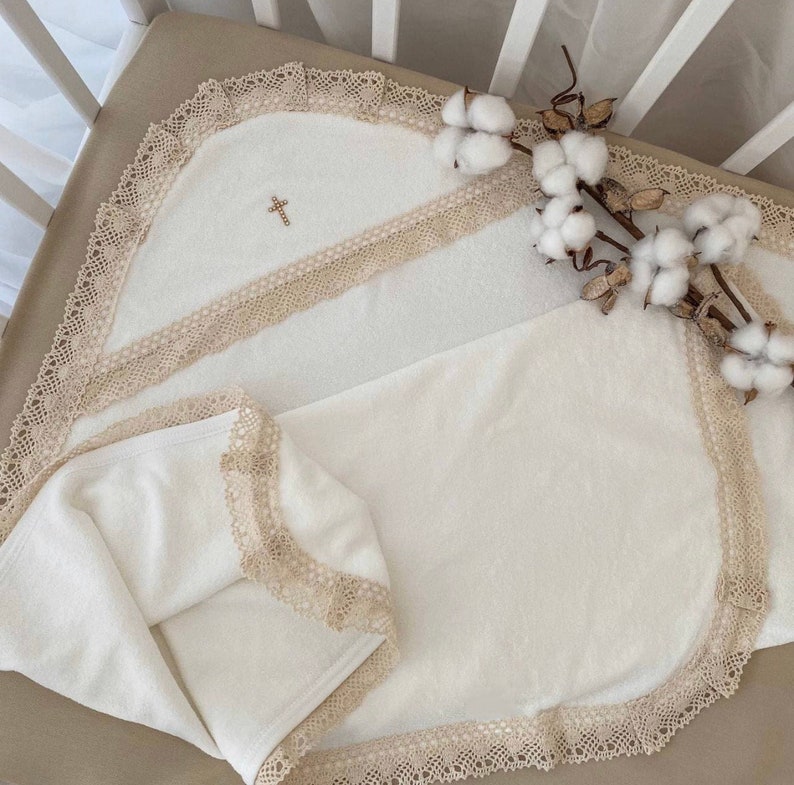 Op de afbeelding: Een wit babydekentje met een geborduurd kruis erop. Het dekentje is afgewerkt met beige kant en ligt op een witte wiegmatras.