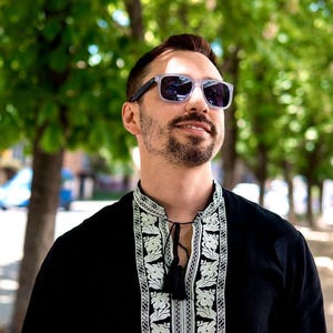 May include: A person wearing clear-framed sunglasses with dark lenses and a black shirt with white floral embroidery. The shirt has a tassel tie at the neckline. The background is blurred with green trees.