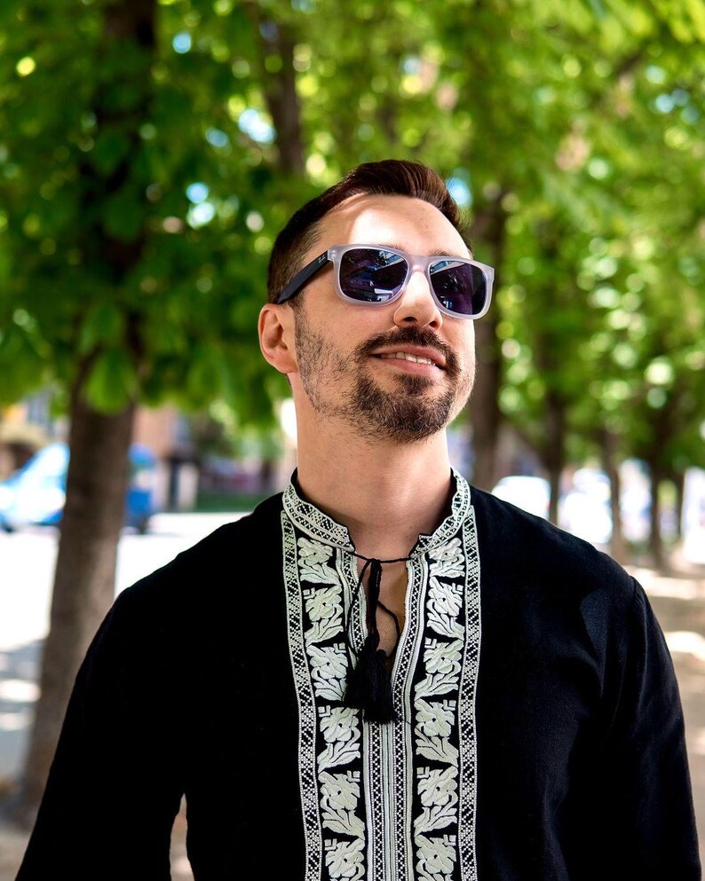 May include: A person wearing clear-framed sunglasses with dark lenses and a black shirt with white floral embroidery. The shirt has a tassel tie at the neckline. The background is blurred with green trees.