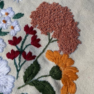 May include: Close-up of embroidered flowers on a cream-colored fabric. The flowers are in shades of red, orange, green, and brown. The flowers are stitched with a variety of techniques, including satin stitch, French knots, and stem stitch.