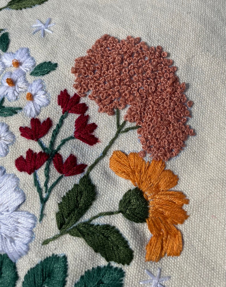 May include: Close-up of embroidered flowers on a cream-colored fabric. The flowers are in shades of red, orange, green, and brown. The flowers are stitched with a variety of techniques, including satin stitch, French knots, and stem stitch.