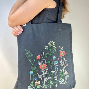 以下が含まれることがあります： 刺繍の花柄が施された黒色のトートバッグ。赤いポピー、青いヤグルマギク、その他のカラフルな野花が緑色の茎と葉で刺繍されています。バッグには黒いハンドルが2つ付いています。