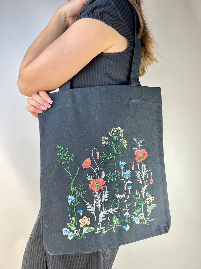 以下が含まれることがあります： 刺繍の花柄が施された黒色のトートバッグ。赤いポピー、青いヤグルマギク、その他のカラフルな野花が緑色の茎と葉で刺繍されています。バッグには黒いハンドルが2つ付いています。