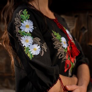 May include: A black embroidered blouse with white flowers and green leaves. The blouse has a red tassel on the neckline.