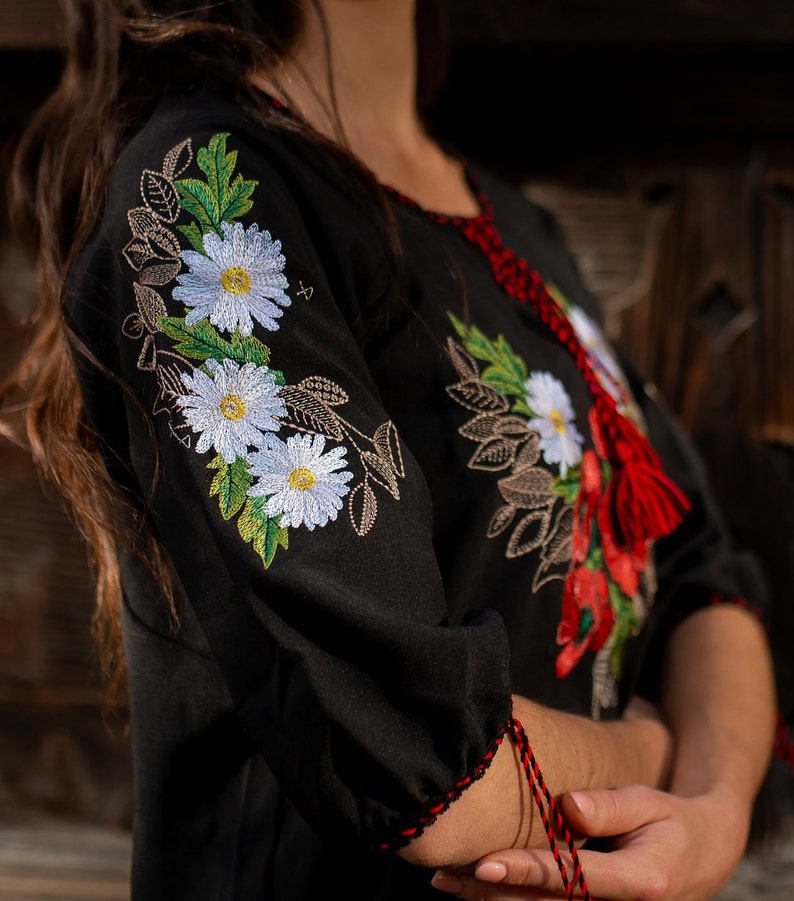 May include: A black embroidered blouse with white flowers and green leaves. The blouse has a red tassel on the neckline.