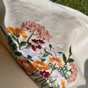 May include: A white canvas tote bag with embroidered flowers in shades of red, orange, yellow, white, and green. The flowers are arranged in a cluster and are surrounded by green leaves.