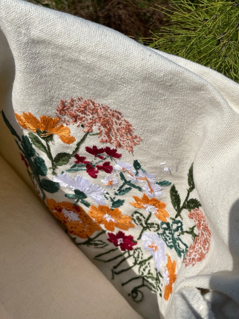 May include: A white canvas tote bag with embroidered flowers in shades of red, orange, yellow, white, and green. The flowers are arranged in a cluster and are surrounded by green leaves.