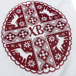 May include: White fabric with a detailed burgundy embroidered design. The design features deer, snowflakes, and geometric patterns within a circular frame. The letters "XB" are also embroidered. The top edge has a fringe and small star-shaped designs.
