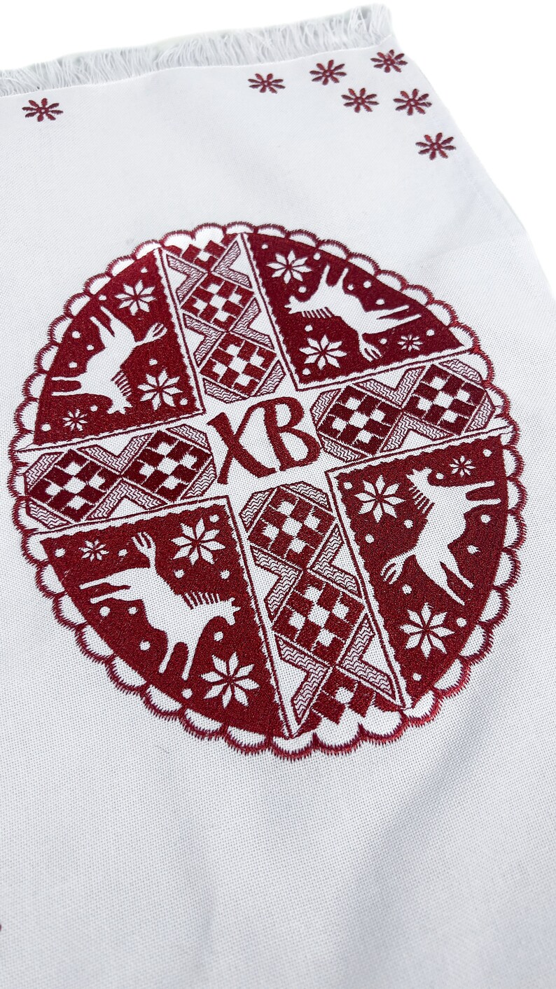 May include: White fabric with a detailed burgundy embroidered design. The design features deer, snowflakes, and geometric patterns within a circular frame. The letters "XB" are also embroidered. The top edge has a fringe and small star-shaped designs.