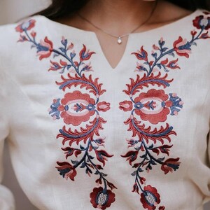 May include: Off-white blouse with detailed floral embroidery in red, blue, and pink. The embroidery is symmetrical, with stylized flowers and leaves. The blouse has a V-neck and long sleeves, suitable for casual or semi-formal wear. The fabric appears to be linen.