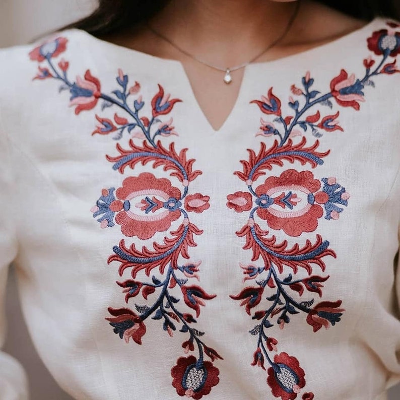 May include: Off-white blouse with detailed floral embroidery in red, blue, and pink. The embroidery is symmetrical, with stylized flowers and leaves. The blouse has a V-neck and long sleeves, suitable for casual or semi-formal wear. The fabric appears to be linen.