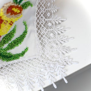 May include: Close-up of a white embroidered textile with a floral design. The embroidery features yellow, orange, and green beads. The edge is finished with a delicate white lace trim, creating a handcrafted look.