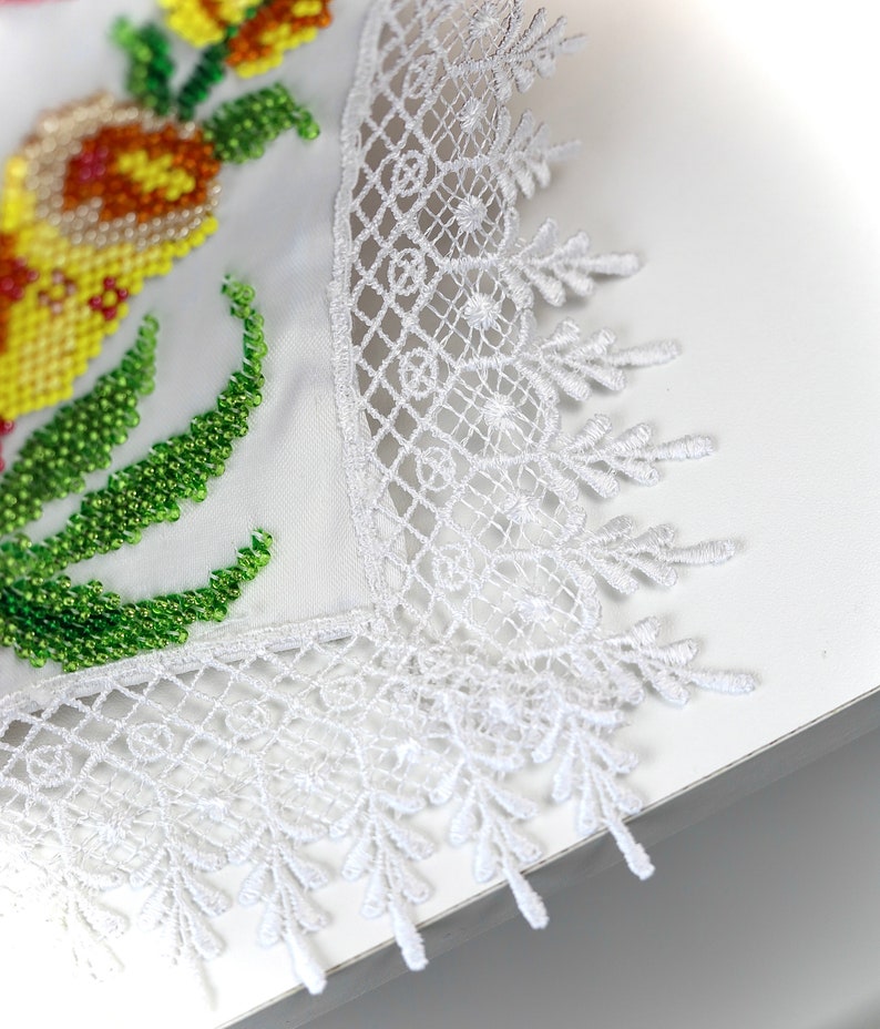 May include: Close-up of a white embroidered textile with a floral design. The embroidery features yellow, orange, and green beads. The edge is finished with a delicate white lace trim, creating a handcrafted look.