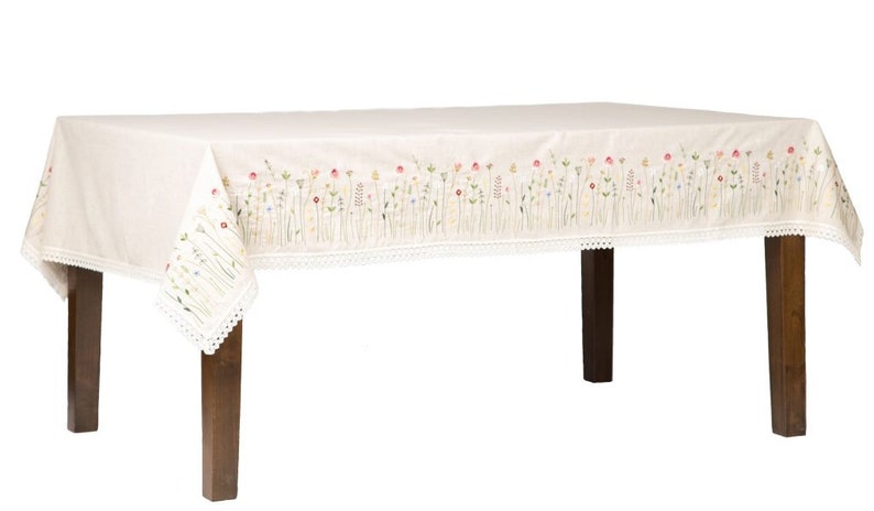 May include: A rectangular cream-colored tablecloth with embroidered floral designs along the edges. The embroidery features colorful flowers and green stems. The tablecloth has a white lace trim and is draped over a wooden table.