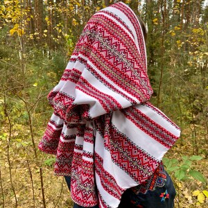 May include: A white headscarf with red and gold geometric embroidery. The scarf is draped over the head and shoulders, with the detailed pattern visible. The background is a blurred forest scene.