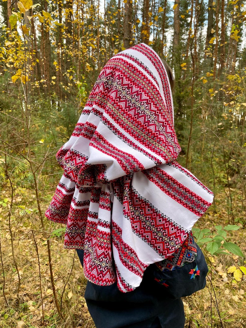 May include: A white headscarf with red and gold geometric embroidery. The scarf is draped over the head and shoulders, with the detailed pattern visible. The background is a blurred forest scene.