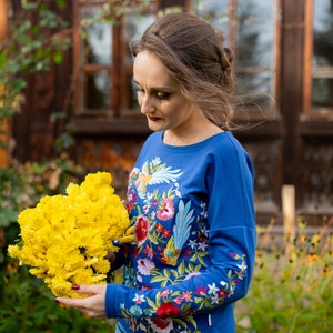 Puede incluir: Una mujer con una camisa azul de manga larga con bordados florales coloridos sostiene un ramo de flores amarillas.