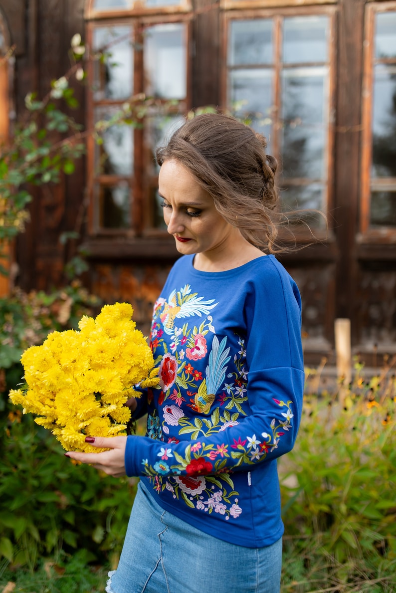 Puede incluir: Una mujer con una camisa azul de manga larga con bordados florales coloridos sostiene un ramo de flores amarillas.