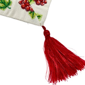 May include: Cream-colored fabric with embroidered green leaves and red grapes. A long, red tassel hangs from the corner. The embroidery details a grapevine design, perfect for home decor.