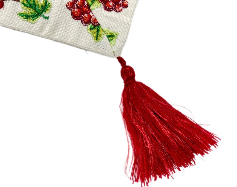 May include: Cream-colored fabric with embroidered green leaves and red grapes. A long, red tassel hangs from the corner. The embroidery details a grapevine design, perfect for home decor.