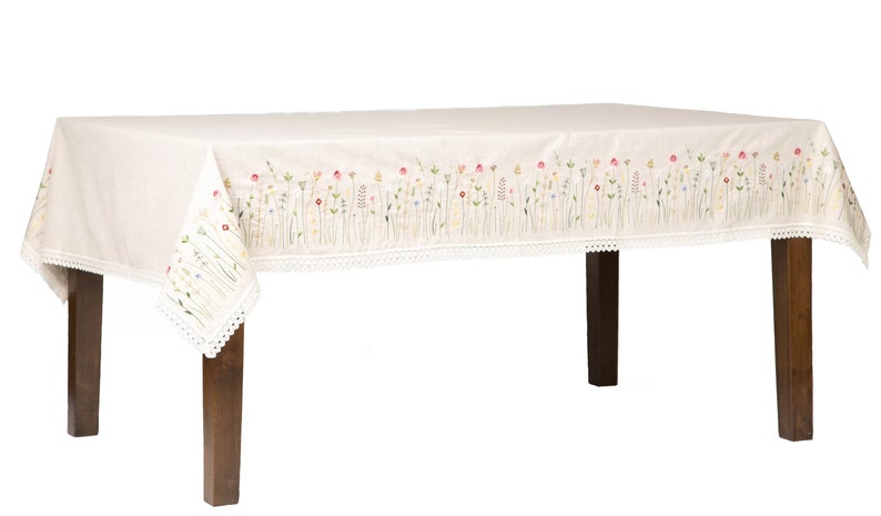 May include: Cream-colored rectangular tablecloth with embroidered floral design. The border features a delicate white lace trim. The floral embroidery includes red, yellow, and purple flowers. The tablecloth is draped over a dark wooden table.