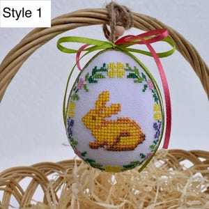 Op de afbeelding: Witte paasei-ornament met een geel kruissteekkonijnontwerp, omgeven door een bloemenkrans. Het ei wordt opgehangen door een touwlus en kleurrijke linten, gepresenteerd in een rieten mand. De tekst "Style 1" staat linksboven.