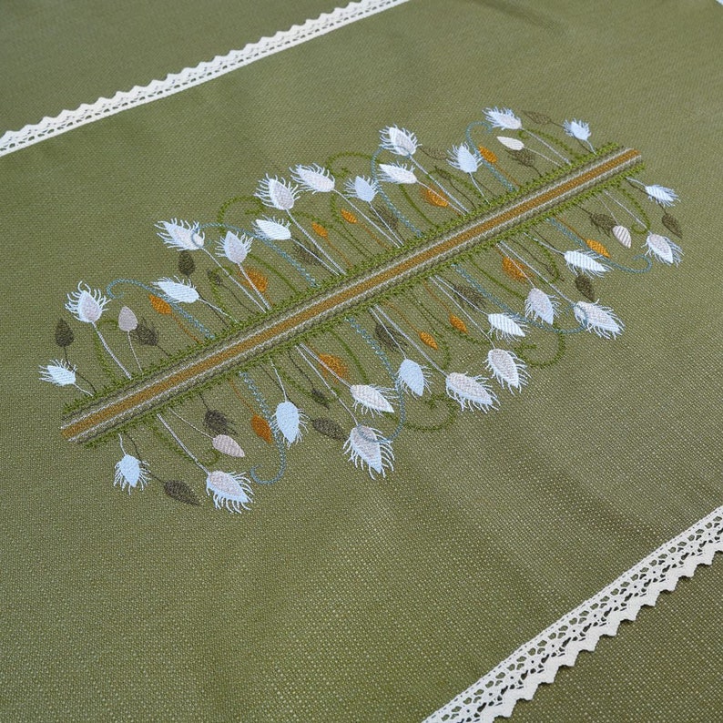 以下が含まれることがあります： オリーブグリーンのテーブルランナー。刺繍された花柄のデザインで、中央のデザインは白、タン、茶色の花の要素が特徴です。端は白いレースでトリミングされており、この織物に繊細さを加えています。