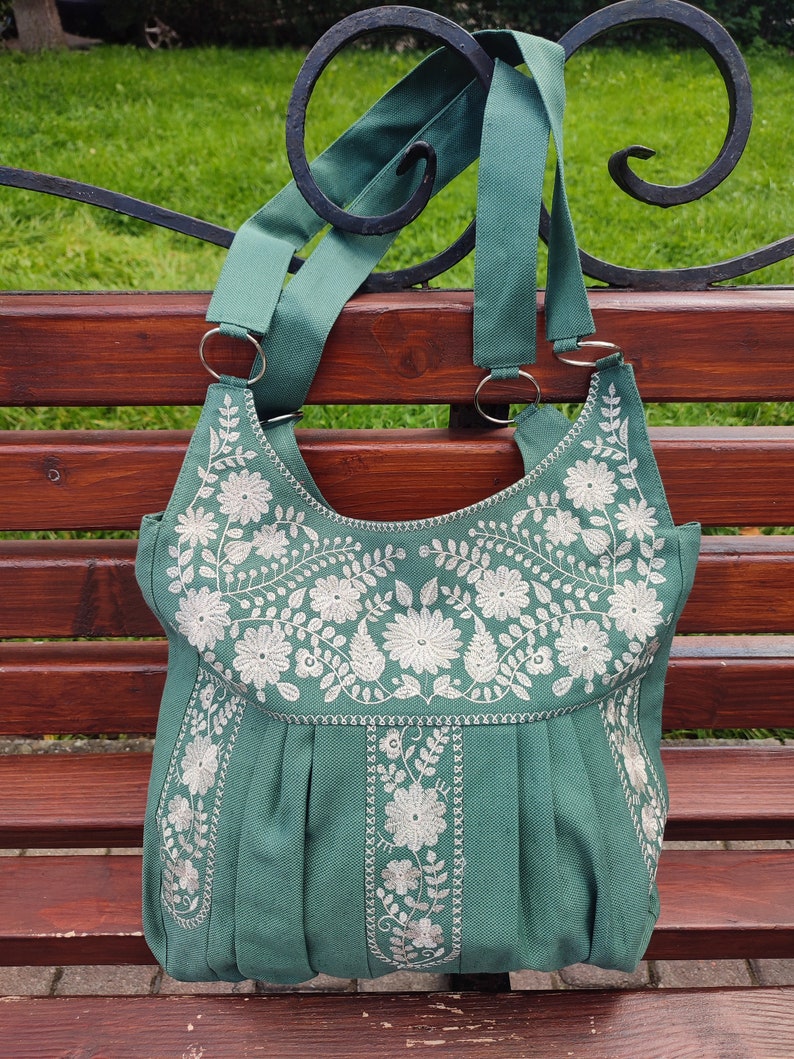 Peut inclure: Un sac cabas en toile vert sarcelle avec des motifs floraux brod&eacute;s blancs. Le sac a deux longues anses et un fond pliss&eacute;.