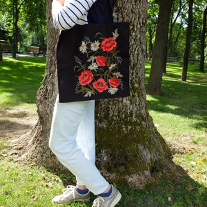 Op de afbeelding: Een zwarte tote bag met geborduurde rode papavers en witte madeliefjes. De tas wordt gedragen door iemand die een witte broek en een gestreept shirt draagt.