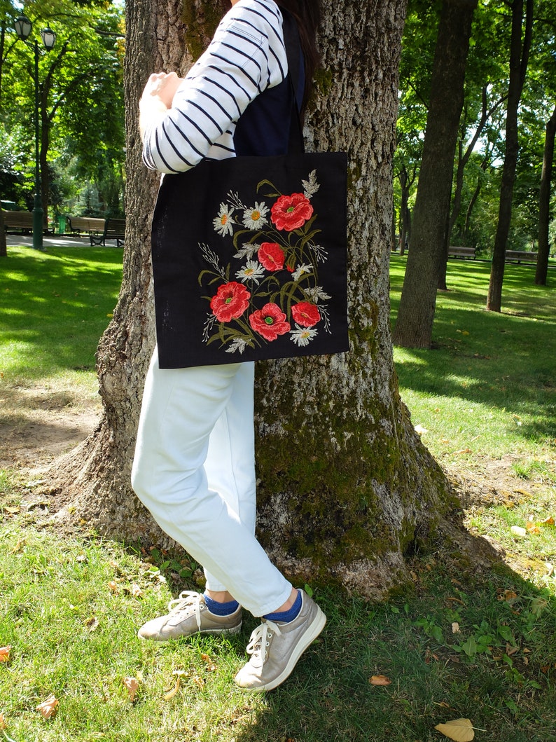 Op de afbeelding: Een zwarte tote bag met geborduurde rode papavers en witte madeliefjes. De tas wordt gedragen door iemand die een witte broek en een gestreept shirt draagt.