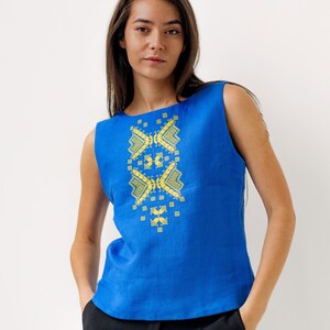 May include: A sleeveless blue linen top with a yellow embroidered geometric design on the front. The top is being worn by a person with long brown hair.