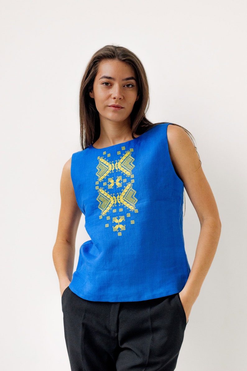 May include: A sleeveless blue linen top with a yellow embroidered geometric design on the front. The top is being worn by a person with long brown hair.