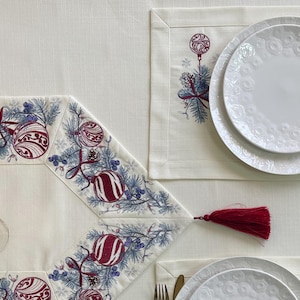 Puede incluir: Una mesa festiva con un mantel crema, con un camino de mesa y manteles individuales con motivos navide&ntilde;os. El camino de mesa y los manteles individuales est&aacute;n adornados con adornos bordados, ramas de pino y bayas en rojo y azul. Platos blancos y cubiertos dorados completan la elegante exhibici&oacute;n navide&ntilde;a.