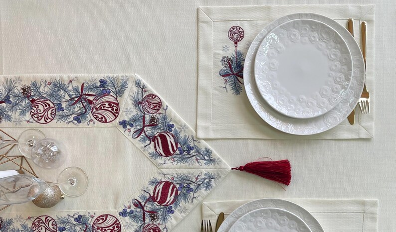 Puede incluir: Una mesa festiva con un mantel crema, con un camino de mesa y manteles individuales con motivos navide&ntilde;os. El camino de mesa y los manteles individuales est&aacute;n adornados con adornos bordados, ramas de pino y bayas en rojo y azul. Platos blancos y cubiertos dorados completan la elegante exhibici&oacute;n navide&ntilde;a.