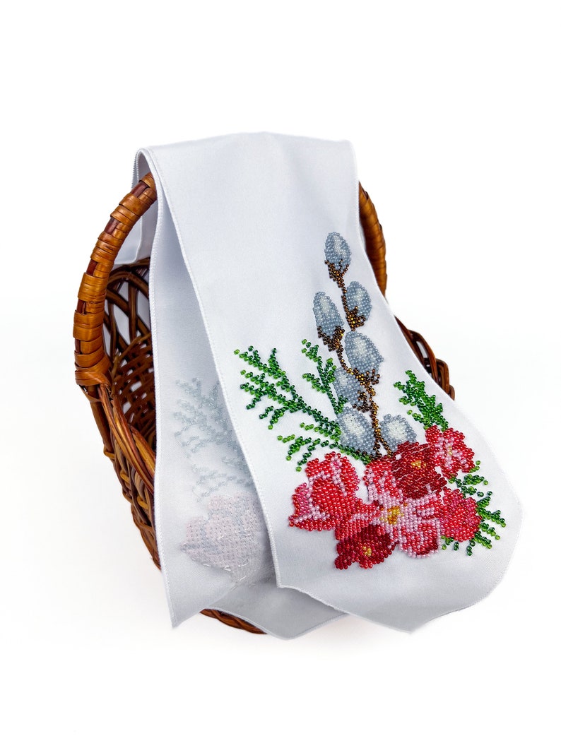 May include: White fabric with a beaded embroidery design of a floral bouquet with red, green, and white flowers and a sprig of willow branches. The fabric is folded and placed in a wicker basket.