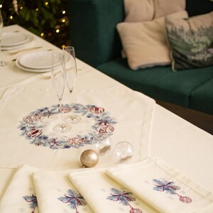 Puede incluir: Una mesa navide&ntilde;a festiva con un mantel y servilletas color crema. La pieza central presenta un dise&ntilde;o de corona con adornos. Las copas de champ&aacute;n y las bolas decorativas se suman al ambiente festivo. Un &aacute;rbol de Navidad y un sof&aacute; verde est&aacute;n en el fondo.