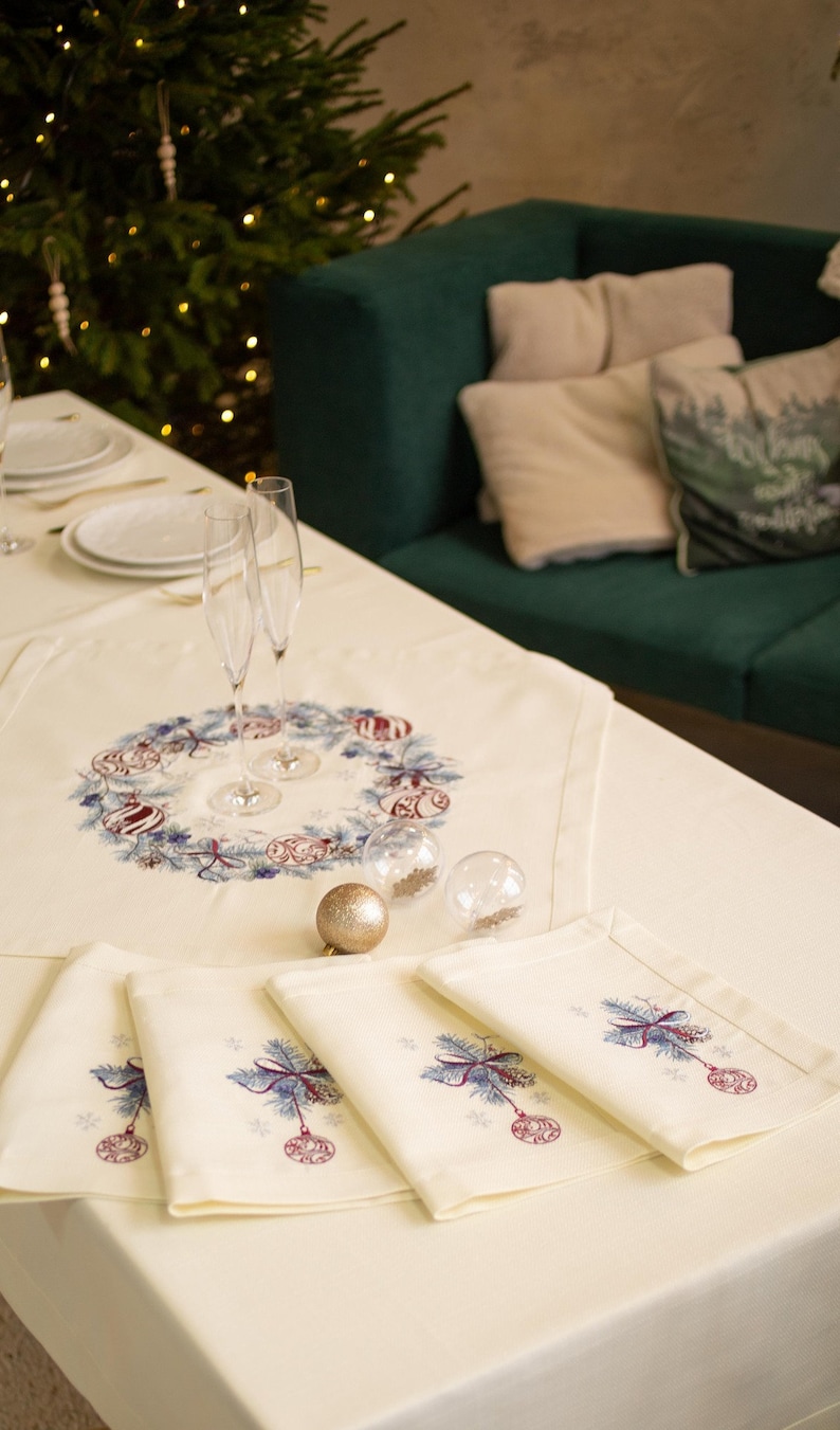 Puede incluir: Una mesa navide&ntilde;a festiva con un mantel y servilletas color crema. La pieza central presenta un dise&ntilde;o de corona con adornos. Las copas de champ&aacute;n y las bolas decorativas se suman al ambiente festivo. Un &aacute;rbol de Navidad y un sof&aacute; verde est&aacute;n en el fondo.