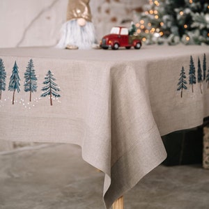 May include: A beige linen tablecloth with a hand-painted design of blue and white pine trees and snow. The tablecloth is draped over a table, with a red toy truck and a gnome figurine in the background. The tablecloth is a festive holiday decoration.