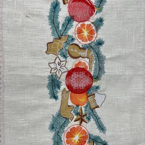 May include: Cream-colored embroidered table runner with a festive design. The design features red ornaments, orange slices, gingerbread cookies, cinnamon sticks, and green pine needles. The runner has a delicate lace trim along the edge.