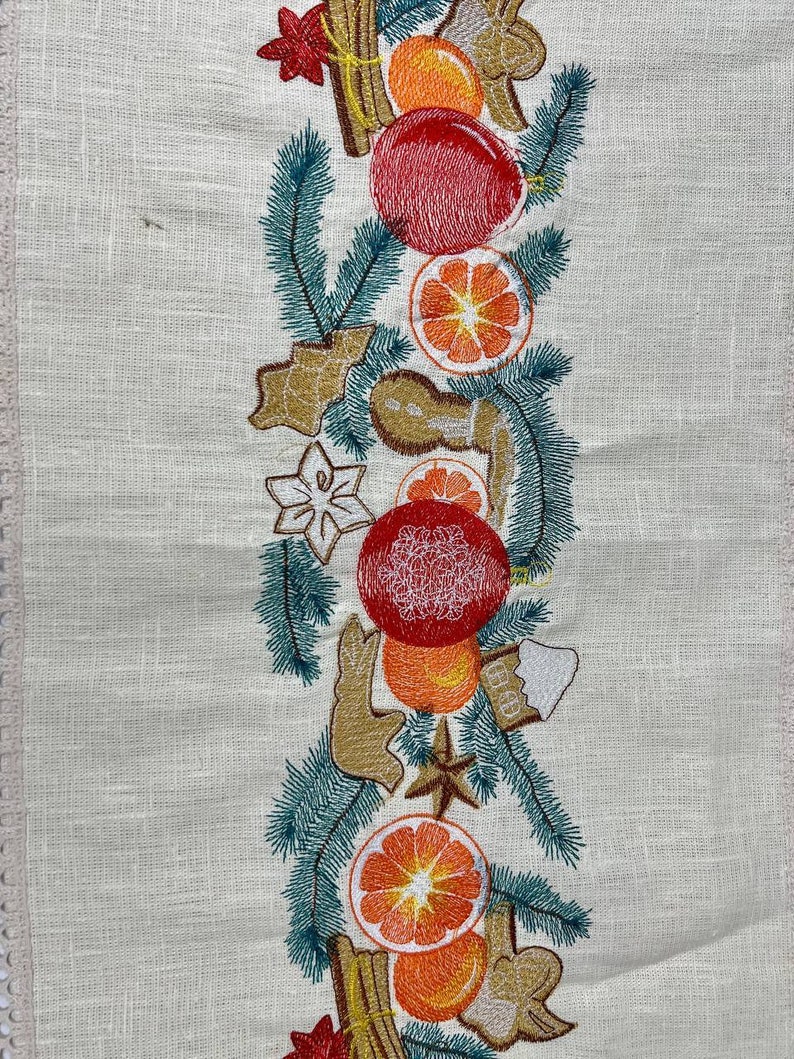 May include: Cream-colored embroidered table runner with a festive design. The design features red ornaments, orange slices, gingerbread cookies, cinnamon sticks, and green pine needles. The runner has a delicate lace trim along the edge.