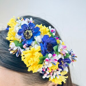 May include: A headband with yellow, blue, and white fabric flowers. The flowers are arranged in a cluster and are attached to a black headband.