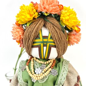Puede incluir: Mu&ntilde;eca hecha a mano con rostro blanco, cabello de hilo marr&oacute;n y una corona floral de flores amarillas y melocot&oacute;n. La mu&ntilde;eca viste un vestido verde con un collar de cuentas de madera y detalles plateados. La cara de la mu&ntilde;eca tiene un dise&ntilde;o geom&eacute;trico.