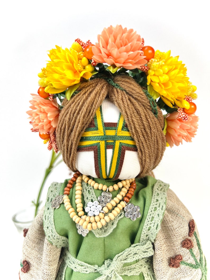 Puede incluir: Mu&ntilde;eca hecha a mano con rostro blanco, cabello de hilo marr&oacute;n y una corona floral de flores amarillas y melocot&oacute;n. La mu&ntilde;eca viste un vestido verde con un collar de cuentas de madera y detalles plateados. La cara de la mu&ntilde;eca tiene un dise&ntilde;o geom&eacute;trico.