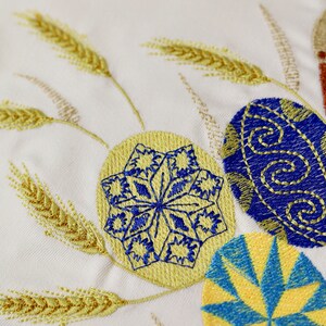 Pu&ograve; includere: Tessuto bianco ricamato con spighe di grano gialle e uova di Pasqua blu e gialle. Le uova hanno diversi motivi, tra cui un fiocco di neve e un design geometrico.