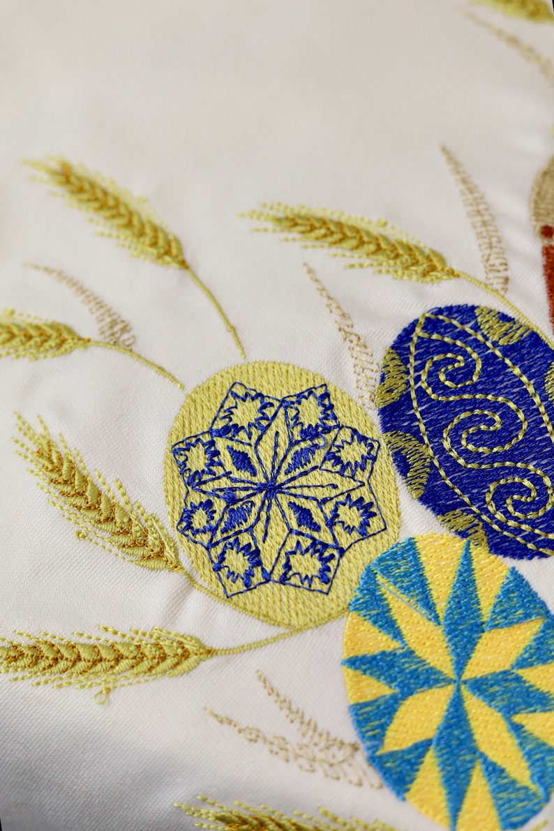 Pu&ograve; includere: Tessuto bianco ricamato con spighe di grano gialle e uova di Pasqua blu e gialle. Le uova hanno diversi motivi, tra cui un fiocco di neve e un design geometrico.