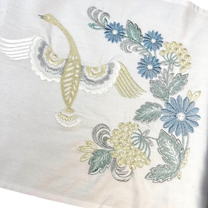 以下が含まれることがあります： 白い生地に、翼を広げた白鳥と花の刺繍が施されています。 白鳥は金、銀、白で、青、緑、黄色の花が咲いています。 刺繍は細かく、デザインは白い背景にあります。