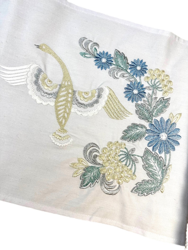 以下が含まれることがあります： 白い生地に、翼を広げた白鳥と花の刺繍が施されています。 白鳥は金、銀、白で、青、緑、黄色の花が咲いています。 刺繍は細かく、デザインは白い背景にあります。