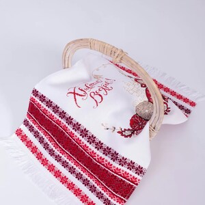Puede incluir: Toalla de lino blanco con bordados florales rojos y burdeos y flecos, expuesta en una cesta de mimbre. La toalla tiene texto bordado en rojo. Un art&iacute;culo decorativo.