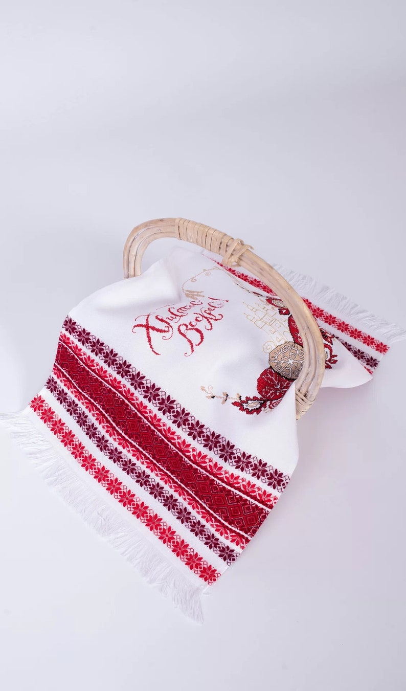 Puede incluir: Toalla de lino blanco con bordados florales rojos y burdeos y flecos, expuesta en una cesta de mimbre. La toalla tiene texto bordado en rojo. Un art&iacute;culo decorativo.